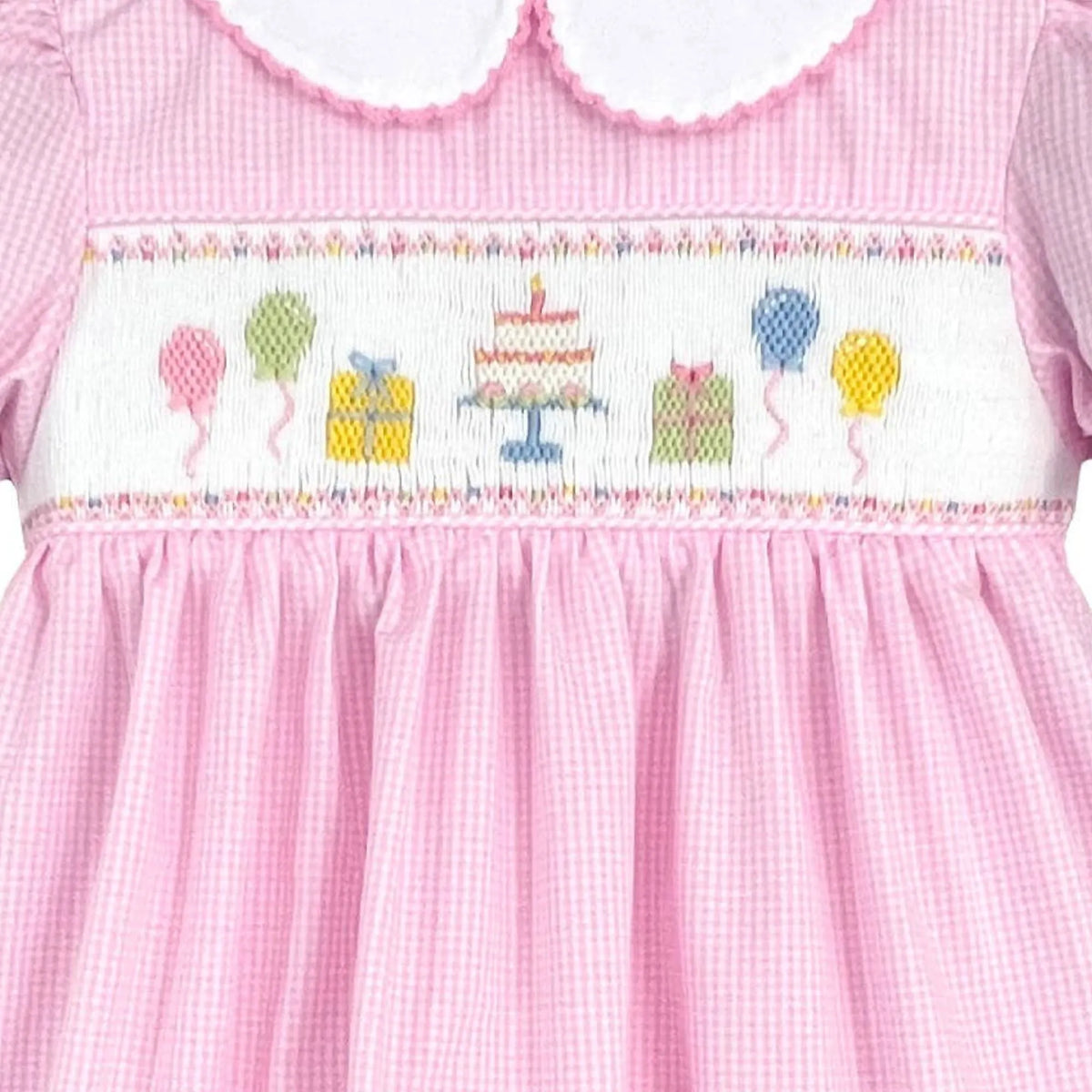 A pink gingham baby dress with a white Peter Pan collar, puffed sleeves, and a smocked chest panel featuring a colorful birthday cake and balloons.